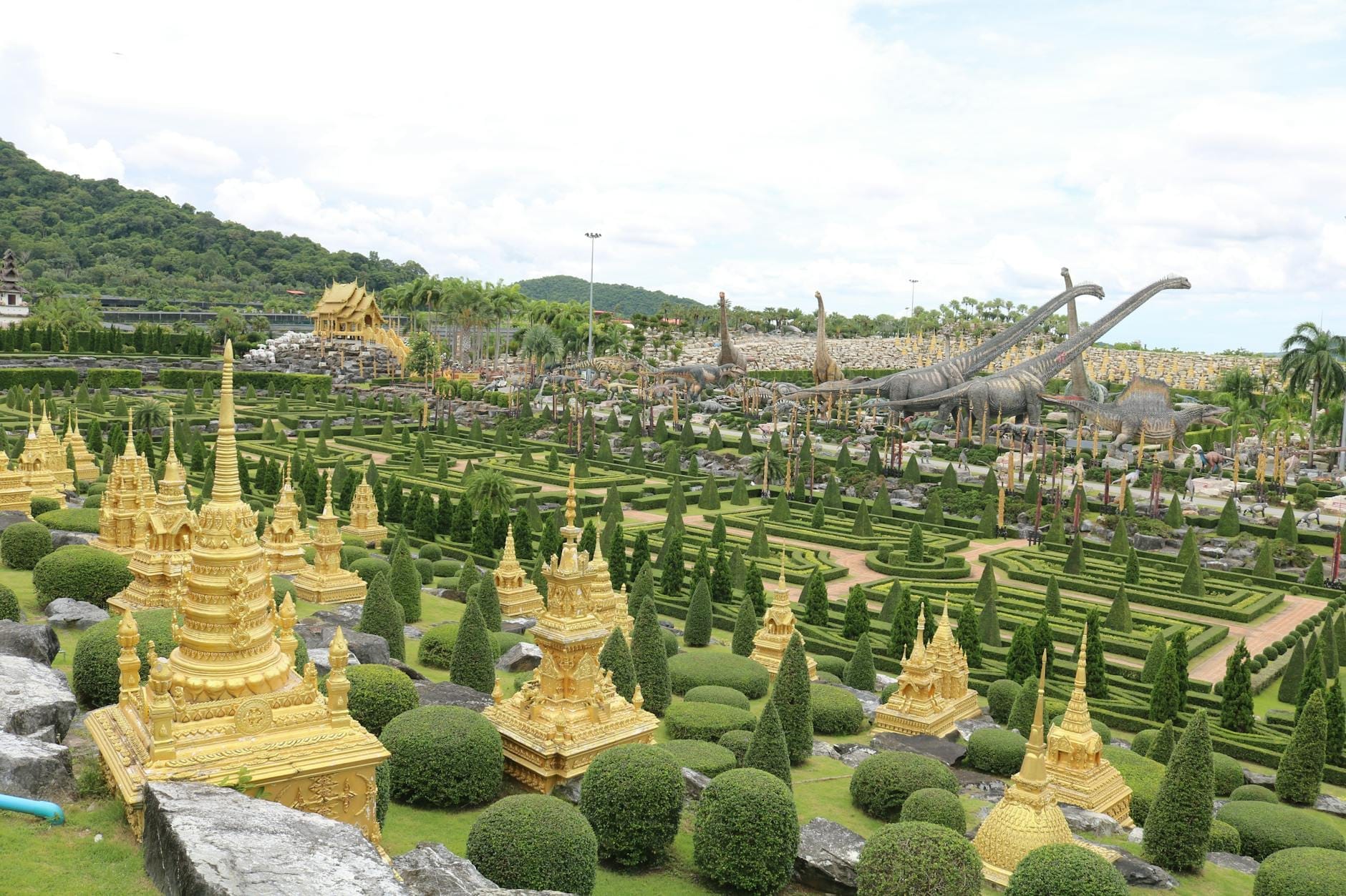 Vista panoramica del Nong Nooch Tropical Garden con stupa dorati, siepi curatissime, alberi topiaria e sagome di dinosauri sullo sfondo sotto un cielo parzialmente nuvoloso.