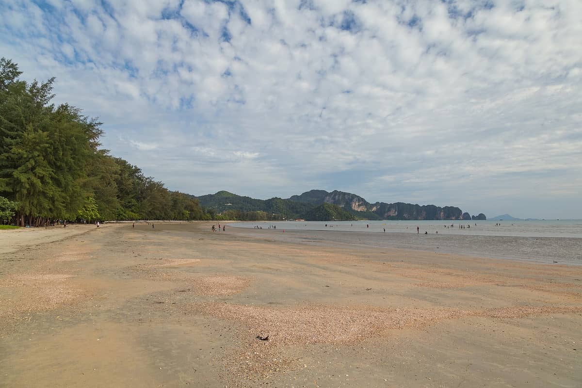 Vista panorámica de la playa Nopparat Thara con orilla de arena, casuarinas a la izquierda, personas caminando y dramáticos acantilados de caliza en la distancia.