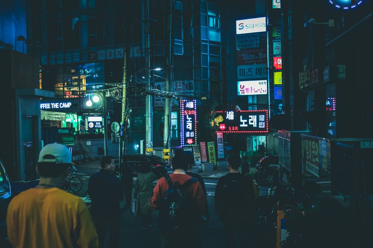 Cena noturna de rua em Seul com letreiros de neon em destaque, incluindo vários com a palavra 'noraebang' em coreano, e pessoas indo em direção aos locais de karaokê.