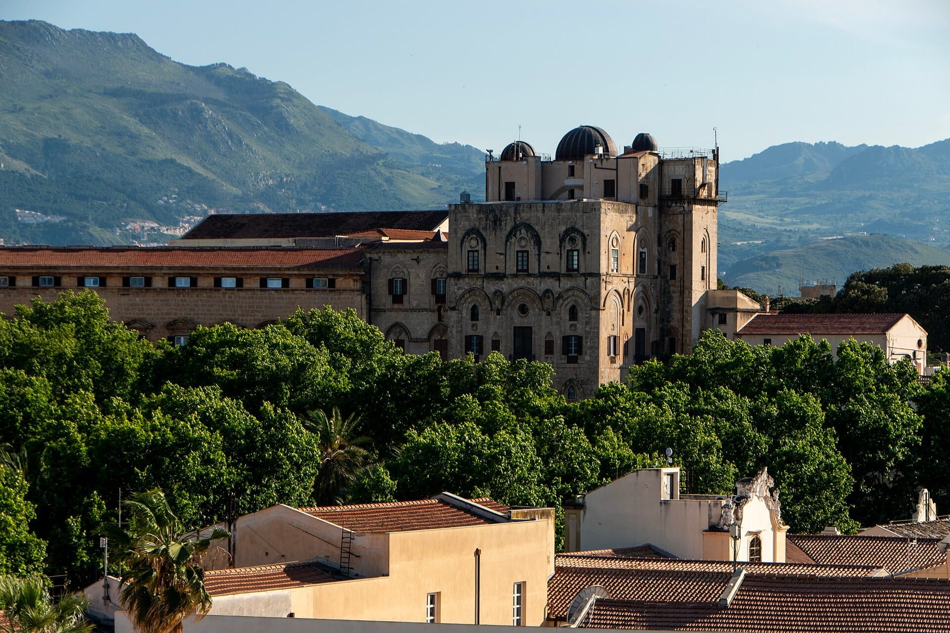 Der Normannische Palast in Palermo steht inmitten üppiger grüner Bäume, mit Berghängen und einem klaren blauen Himmel im Hintergrund.