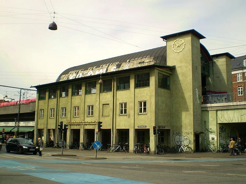วิวถนนของ Nørrebro Station ในโคเปนเฮเกน พร้อมด้วยผนังสีเหลืองอ่อน หลังคาโค้ง นาฬิกาเมือง กราฟิตี จักรยาน และคนเดินเท้าใต้ท้องฟ้าที่มีเมฆมาก