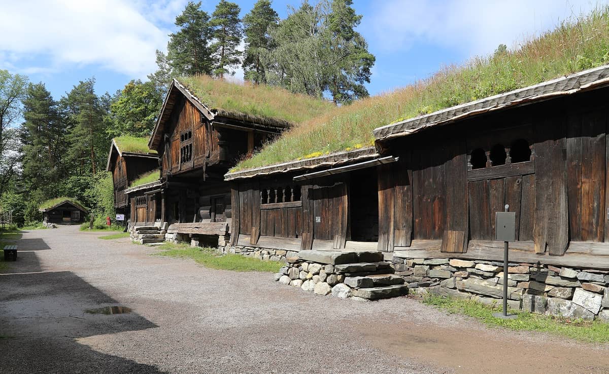 อาคารไม้ซุงแบบดั้งเดิมของนอร์เวย์ที่มีหลังคาหญ้าในพิพิธภัณฑ์ Norsk Folkemuseum ล้อมรอบด้วยต้นไม้ภายใต้ท้องฟ้าสีฟ้าแดดจ้า