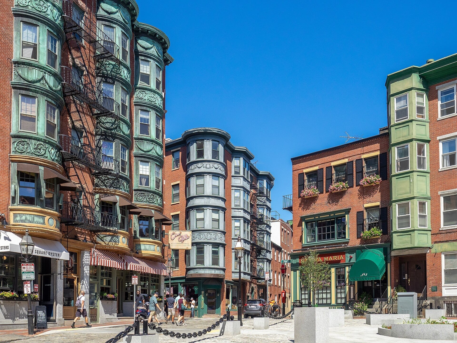 อาคารอิฐและทองแดงทางประวัติศาสตร์เรียงรายตามถนนที่เต็มไปด้วยแสงแดดใน North End เมือง Boston พร้อมผู้คนที่เดินผ่านร้านกาแฟและร้านอาหารใต้ท้องฟ้าสีฟ้าแจ่มใส
