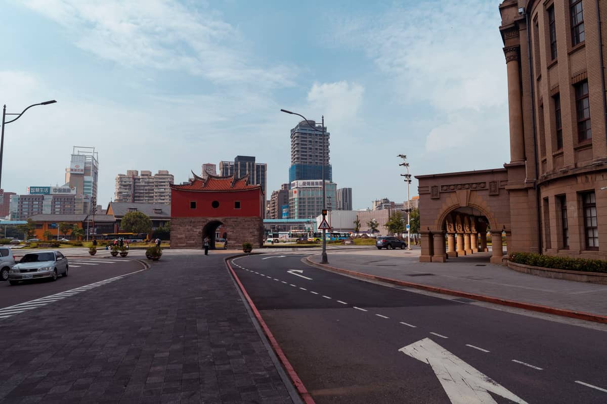 Weitwinkelfoto des Nordtors (Beimen) in Taipeh mit seinem roten Obergeschoss und dem Steinsockel, umgeben von Stadtstraßen und modernen Gebäuden unter blauem Himmel.