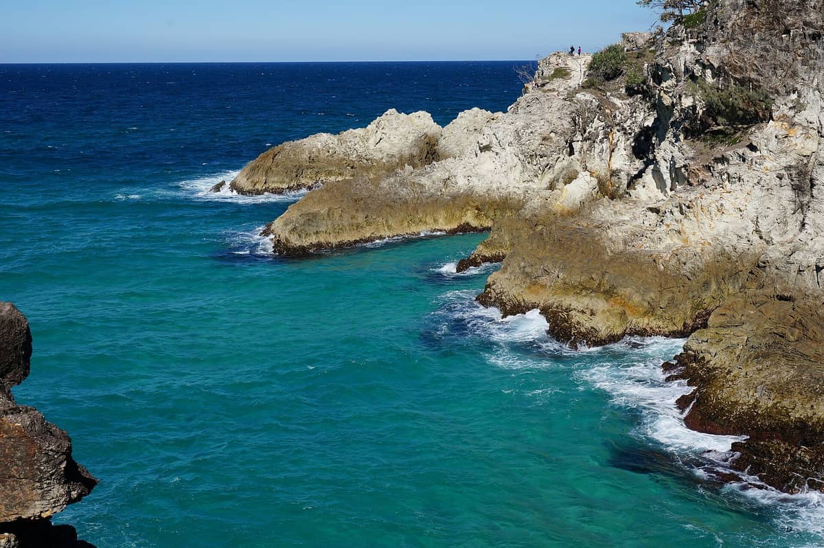 Schroffe Felsenklippen treffen auf türkisfarbene Meereswellen am North Gorge Walk, Point Lookout, unter einem klaren blauen Himmel mit Beobachtern in der Ferne auf dem Vorgebirge.