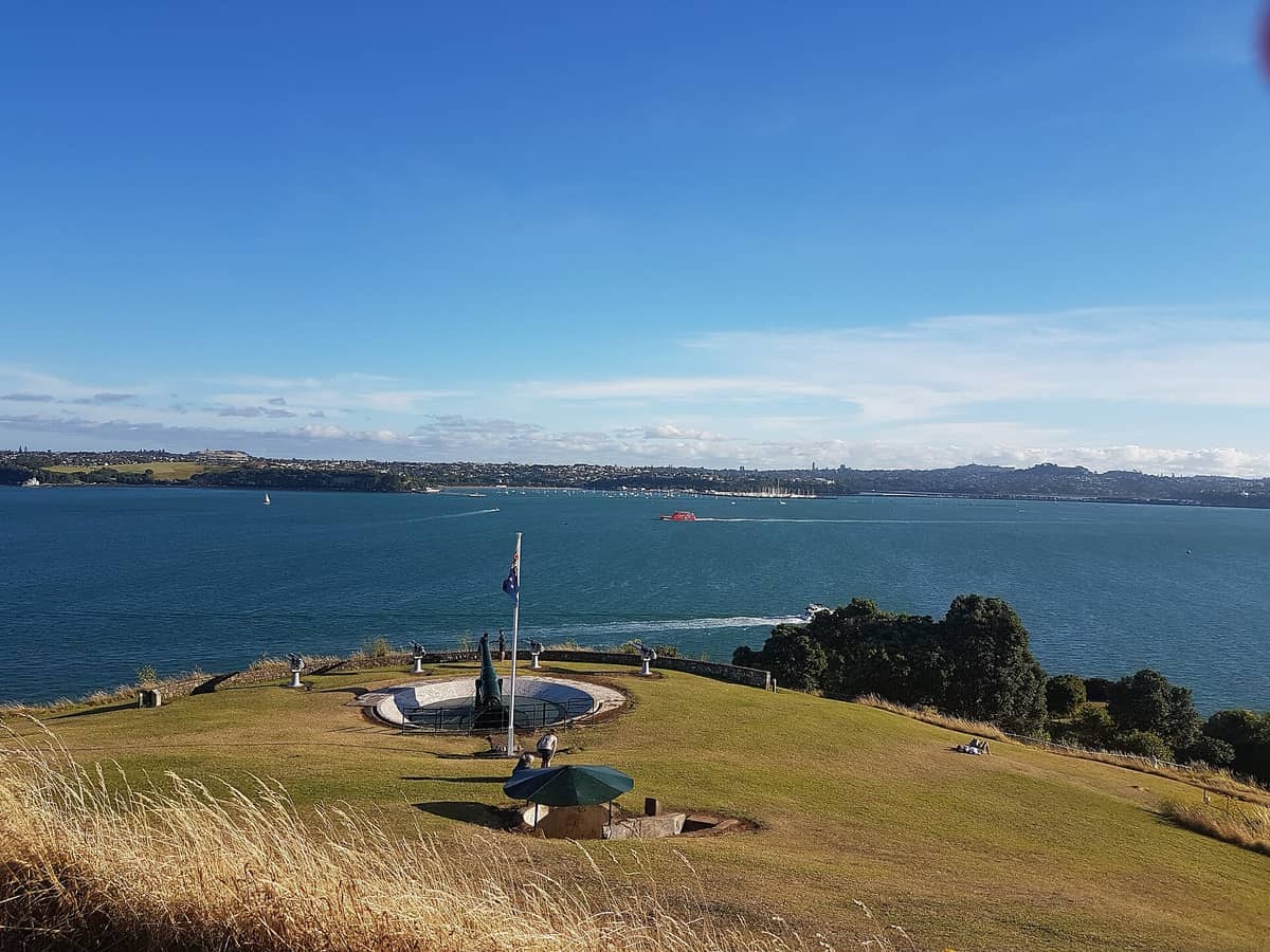 Grasbewachsene Hänge und Geschützstellungen im Maungauika / North Head Historic Reserve mit Blick auf Aucklands Waitematā Harbour unter strahlend blauem Himmel.