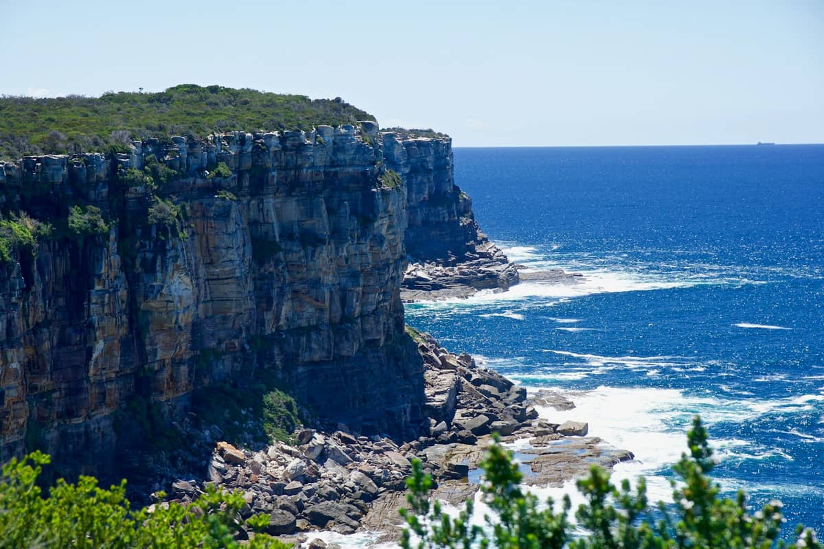 Imponentes acantilados costeros de North Head Sanctuary sobre un océano azul intenso, cubiertos de vegetación autóctona y matorral, bajo un cielo despejado y luz brillante.