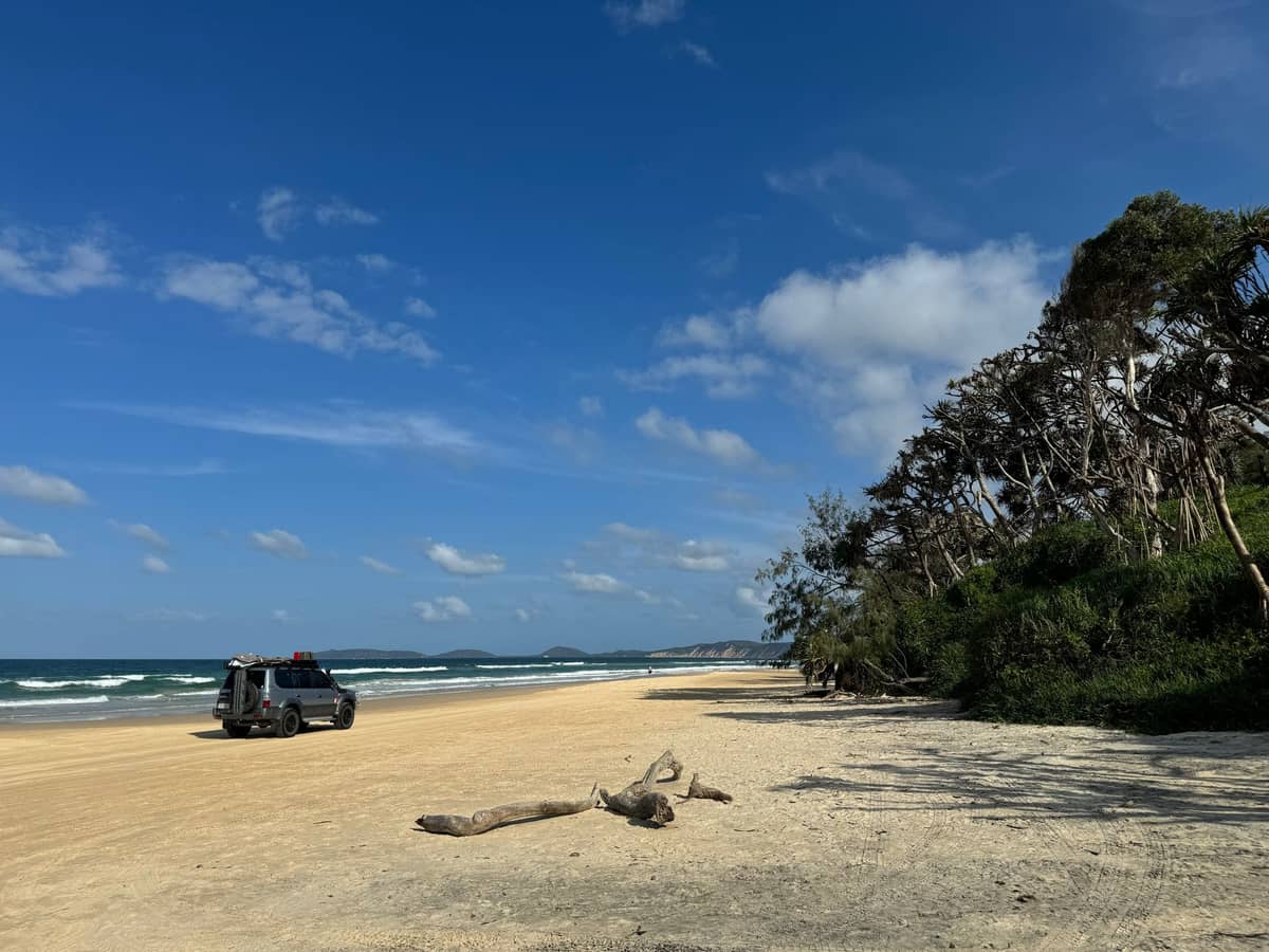 Szeroka, piaszczysta plaża na North Stradbroke Island z samochodem terenowym zaparkowanym przy drzewach, pod błękitnym niebem z lekkimi chmurami i falami dobijającymi do brzegu.