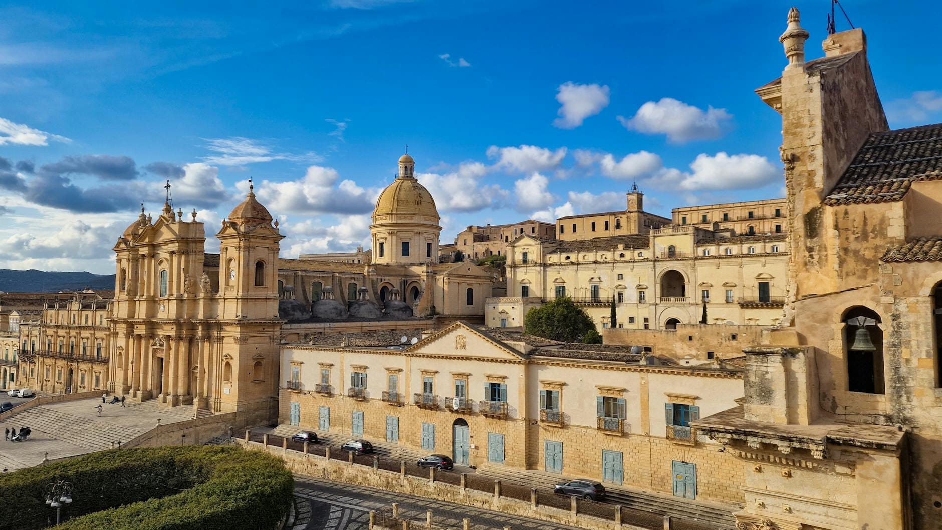 Weitwinkelaufnahme der prächtigen Barockfassade und Kuppel der Noto-Kathedrale, die hoch über ihrer Freitreppe und den umliegenden historischen Gebäuden unter einem leuchtend blauen Himmel aufragt.