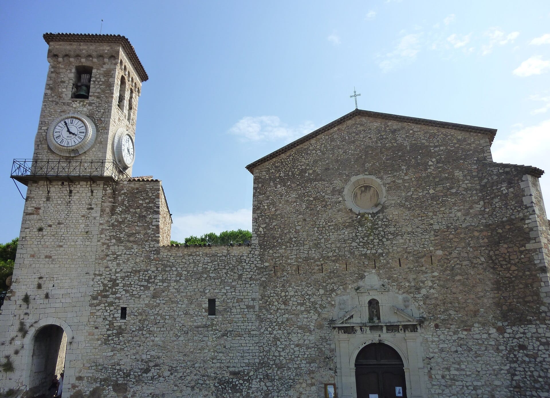 Vista frontale della chiesa Notre-Dame de l'Espérance a Cannes, con le sue storiche mura in pietra e la grande torre dell'orologio sotto un cielo azzurro limpido.