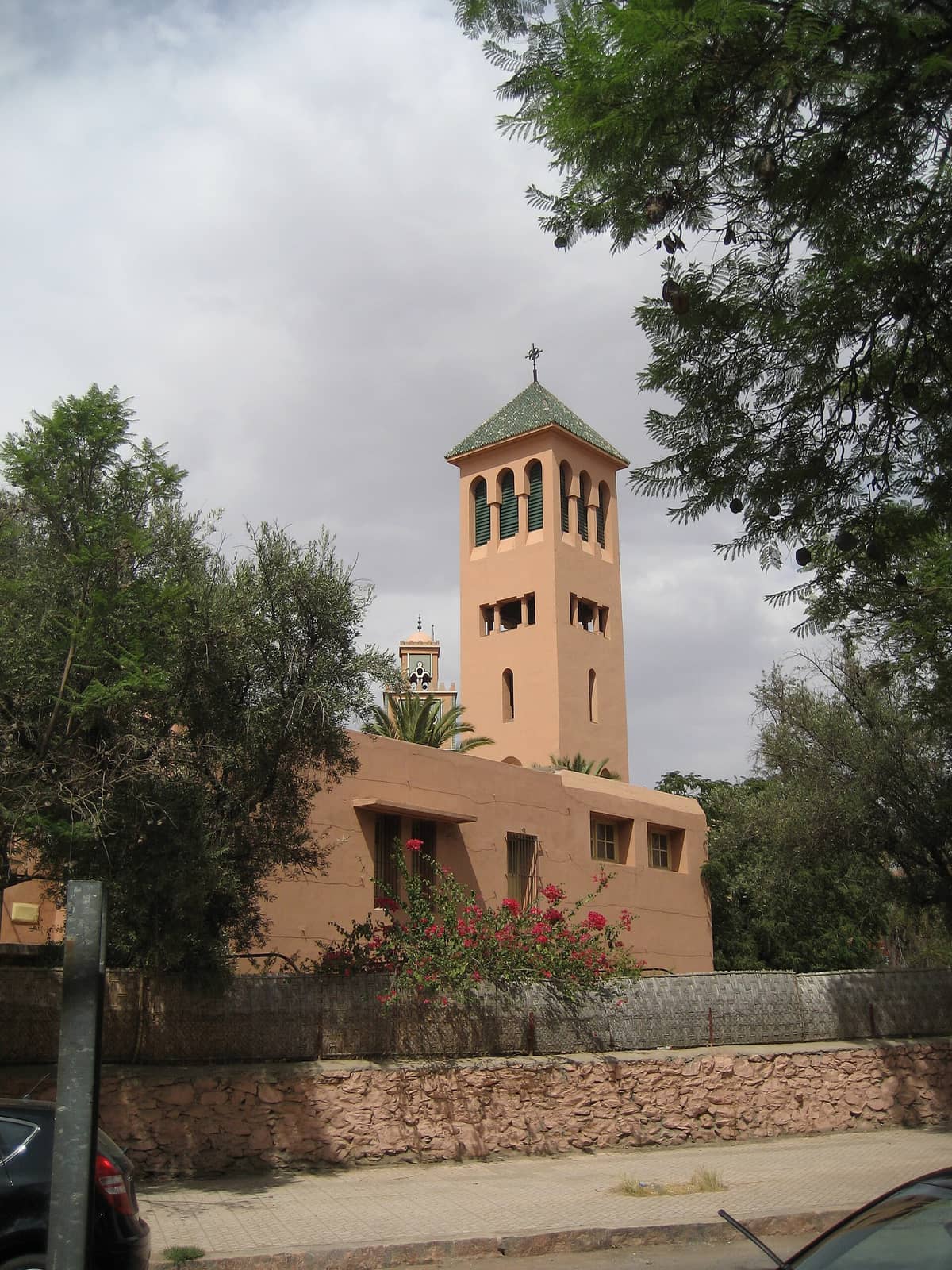 Église des Saints-Martyrs ในย่าน Gueliz ของ Marrakech มีหอโบสถ์สีเบจเรียบง่ายพร้อมไม้กางเขน ล้อมรอบด้วยต้นไม้สีเขียวและท้องฟ้ามีเมฆ