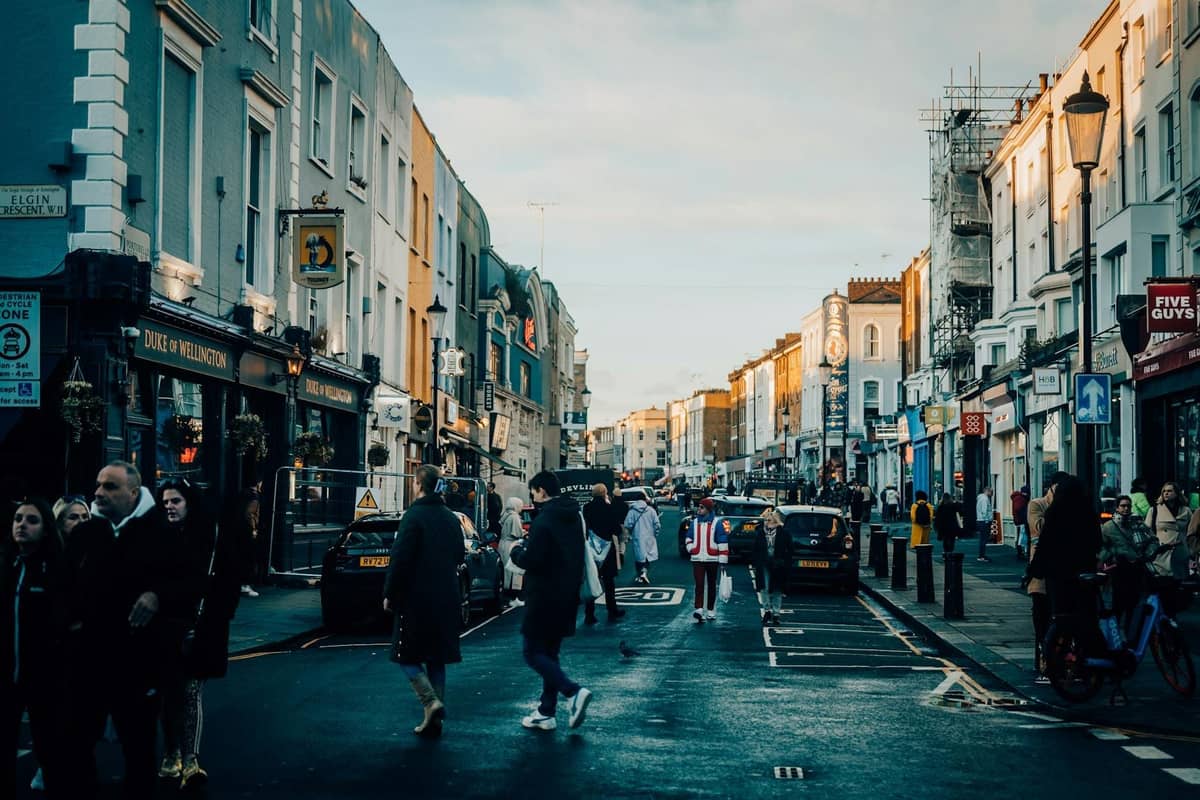 Rua movimentada em Notting Hill com pessoas atravessando, casas coloridas em fila, lojas e arquitetura londrina característica, combinando com a atmosfera da rota do desfile do carnaval.