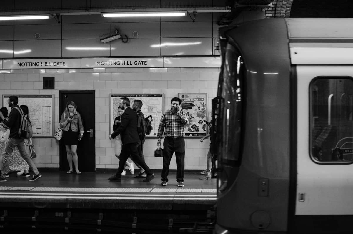 Passageiros em pé e caminhando na plataforma da estação de metrô Notting Hill Gate em Londres com um trem em primeiro plano.