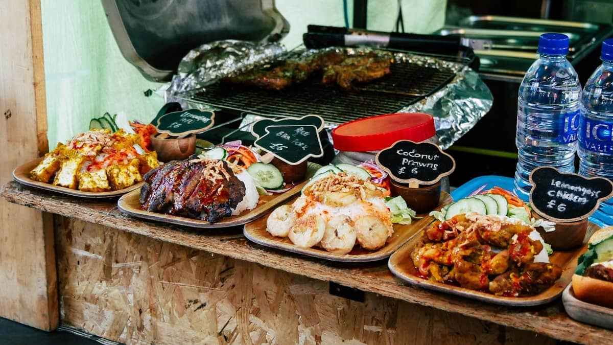 Pratos de comida de rua caribenha com placas de preços escritas à mão e água engarrafada em uma barraca de mercado ao ar livre em Londres.
