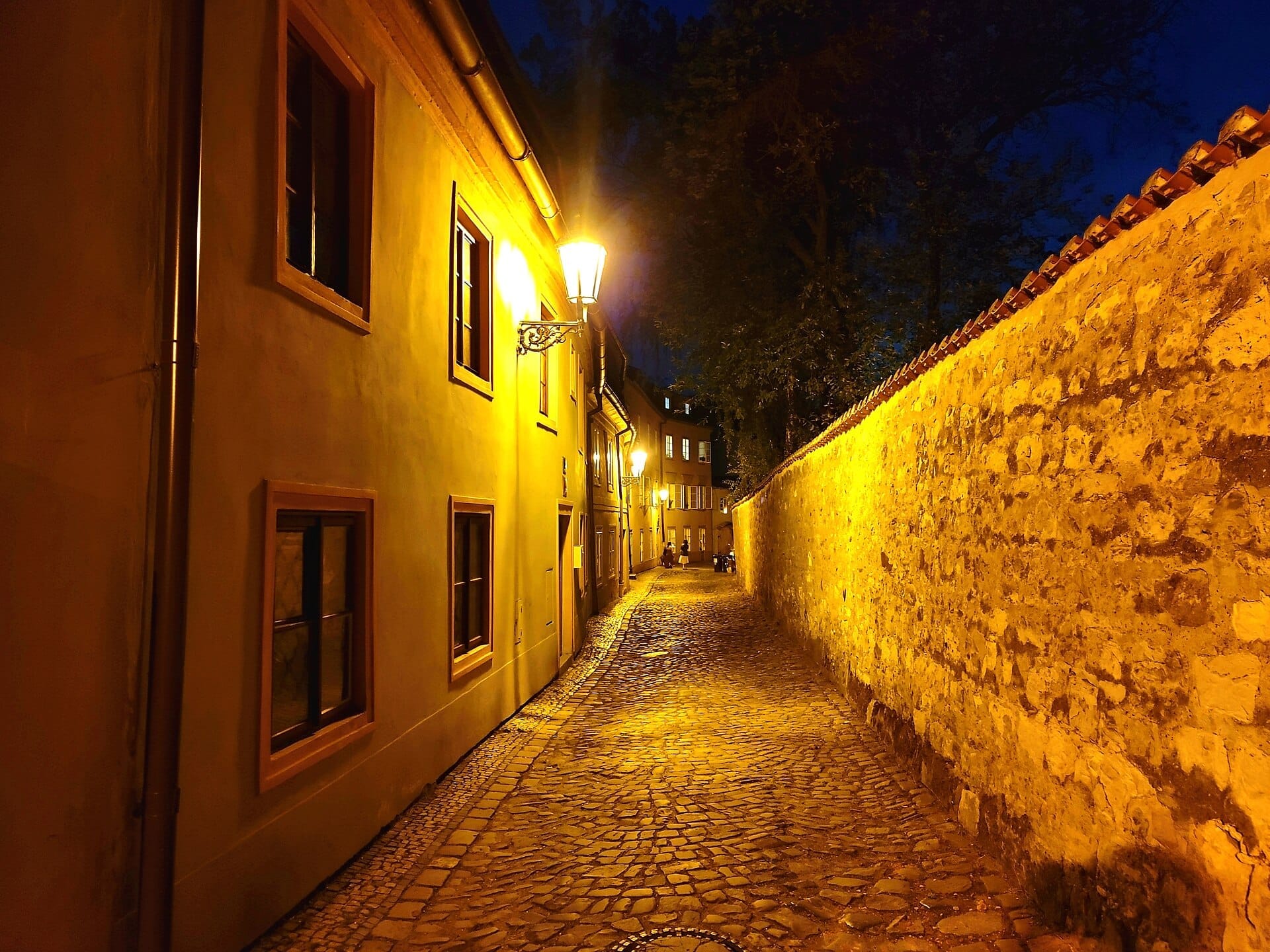 Enge Kopfsteinpflastergasse im Viertel Nový Svět, Prag, gesäumt von historischen Steinmauern und gelblich beleuchteten Gebäuden bei Nacht.