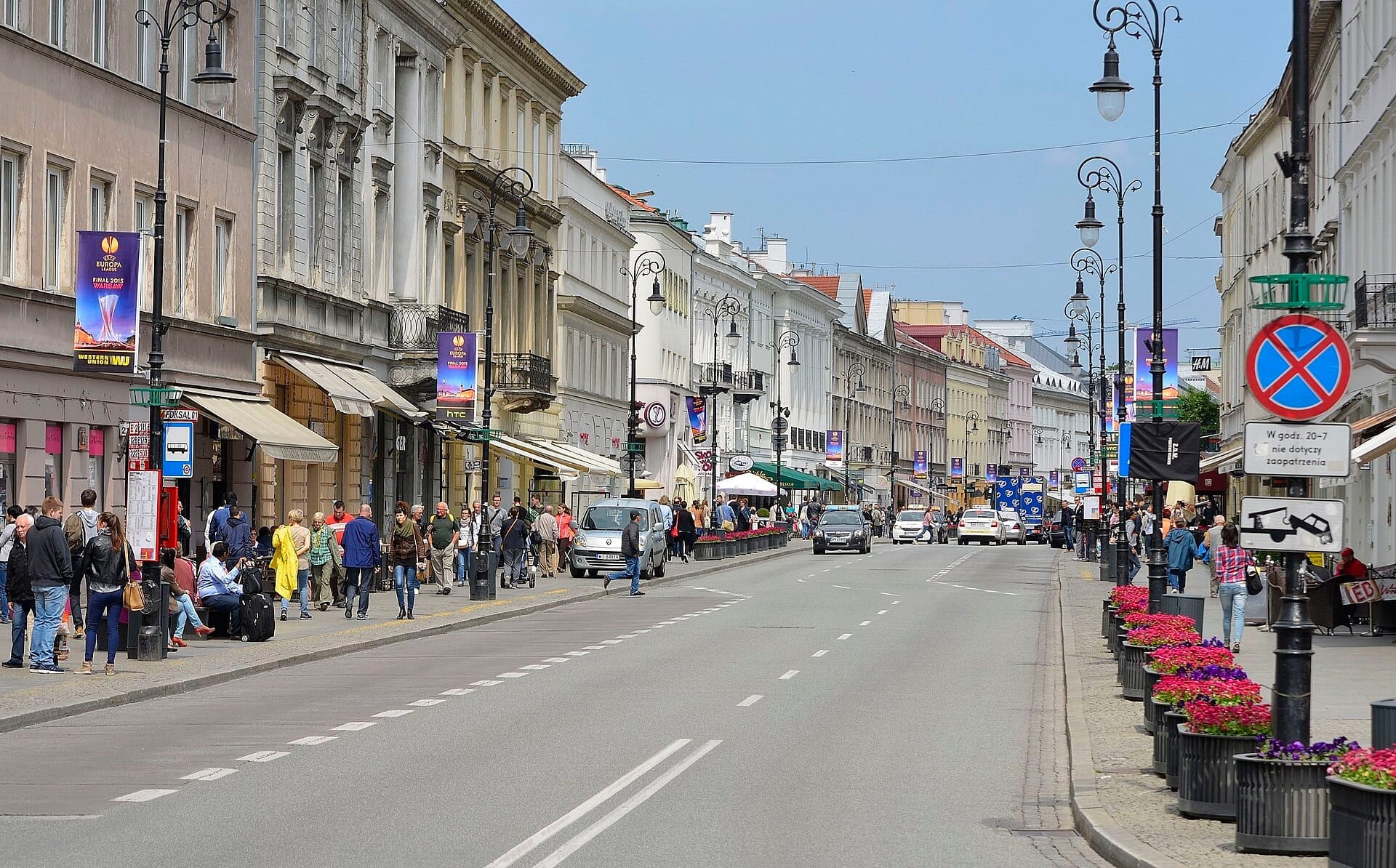 ถนน Nowy Świat ที่คึกคักในกรุง Warsaw มีอาคารสไตล์นีโอคลาสสิก ร้านกาแฟกลางแจ้ง เสาไฟริมถนน และผู้คนเดินชมการจัดแสดงดอกไม้หลากสีสันในวันที่แสงแดดสดใส