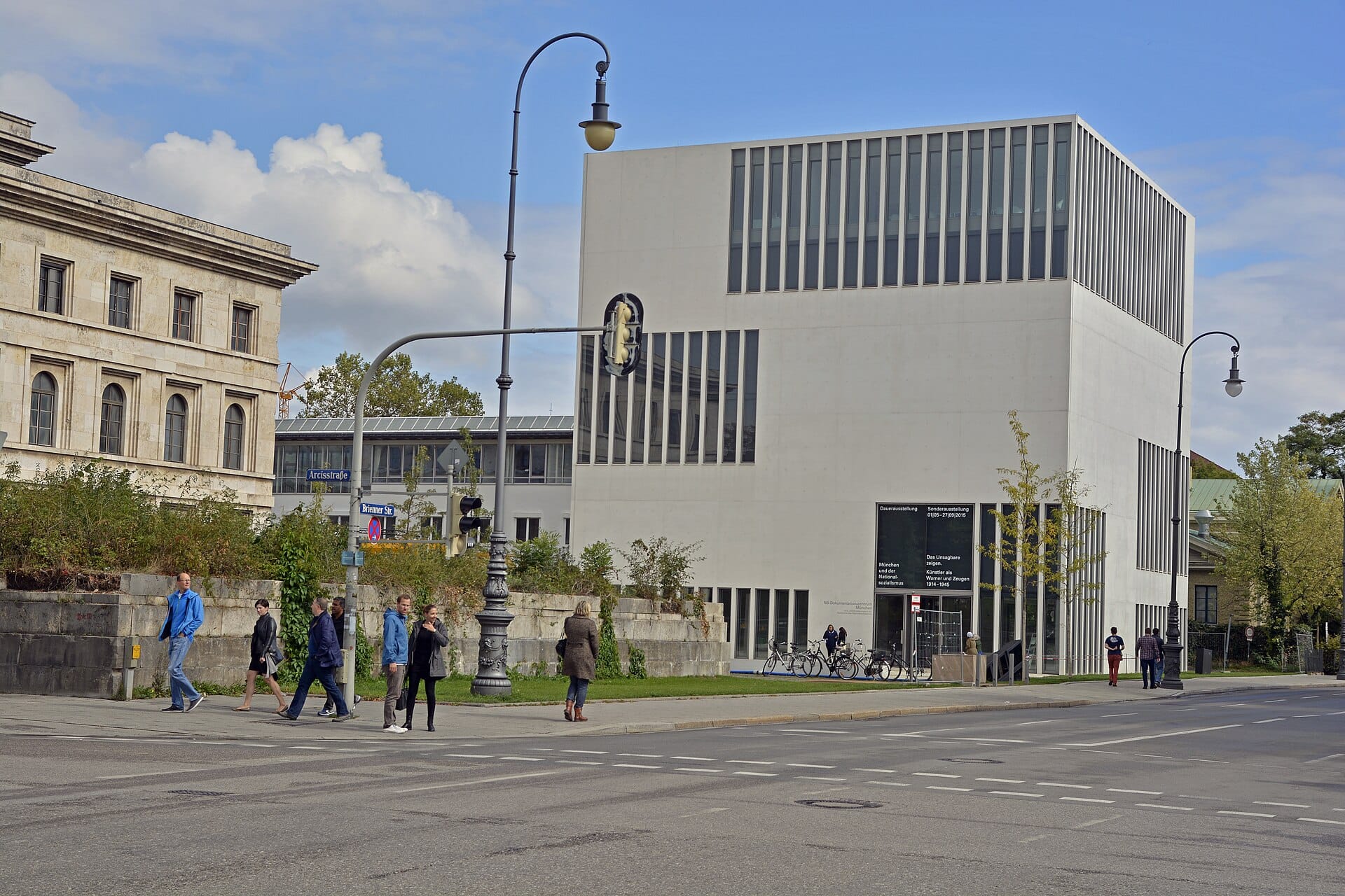 ด้านนอกของ NS-Dokumentationszentrum München อาคารสีขาวสี่เหลี่ยมทันสมัยมีหน้าต่างบานใหญ่ ถ่ายจากอีกฝั่งถนนในวันฟ้าใส