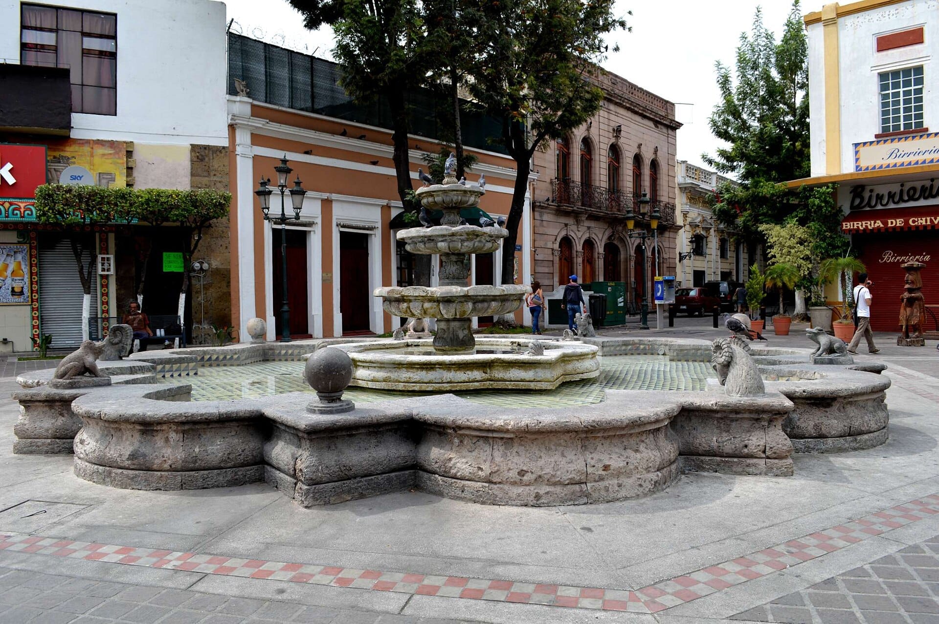Grande fontana in pietra al centro di Plaza Las Nueve Esquinas, circondata da edifici coloniali e alcuni pedoni in una giornata nuvolosa.