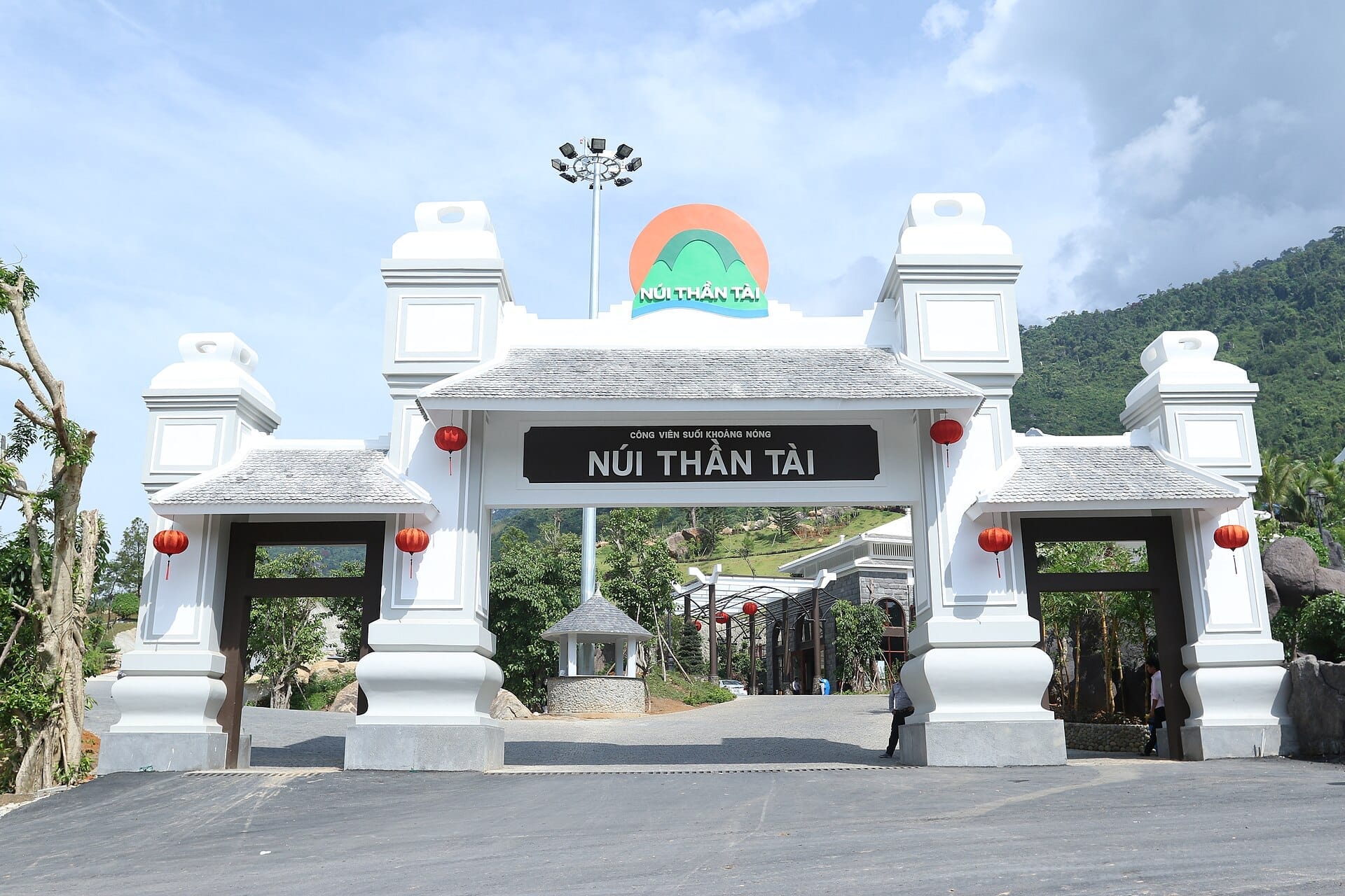ประตูทางเข้าสีขาวขนาดใหญ่ของ Nui Than Tai Hot Springs Park ที่ประดับด้วยโคมไฟสีแดง ตั้งตระหง่านอยู่เบื้องหน้าภูเขาสีเขียวขจีอันอุดมสมบูรณ์