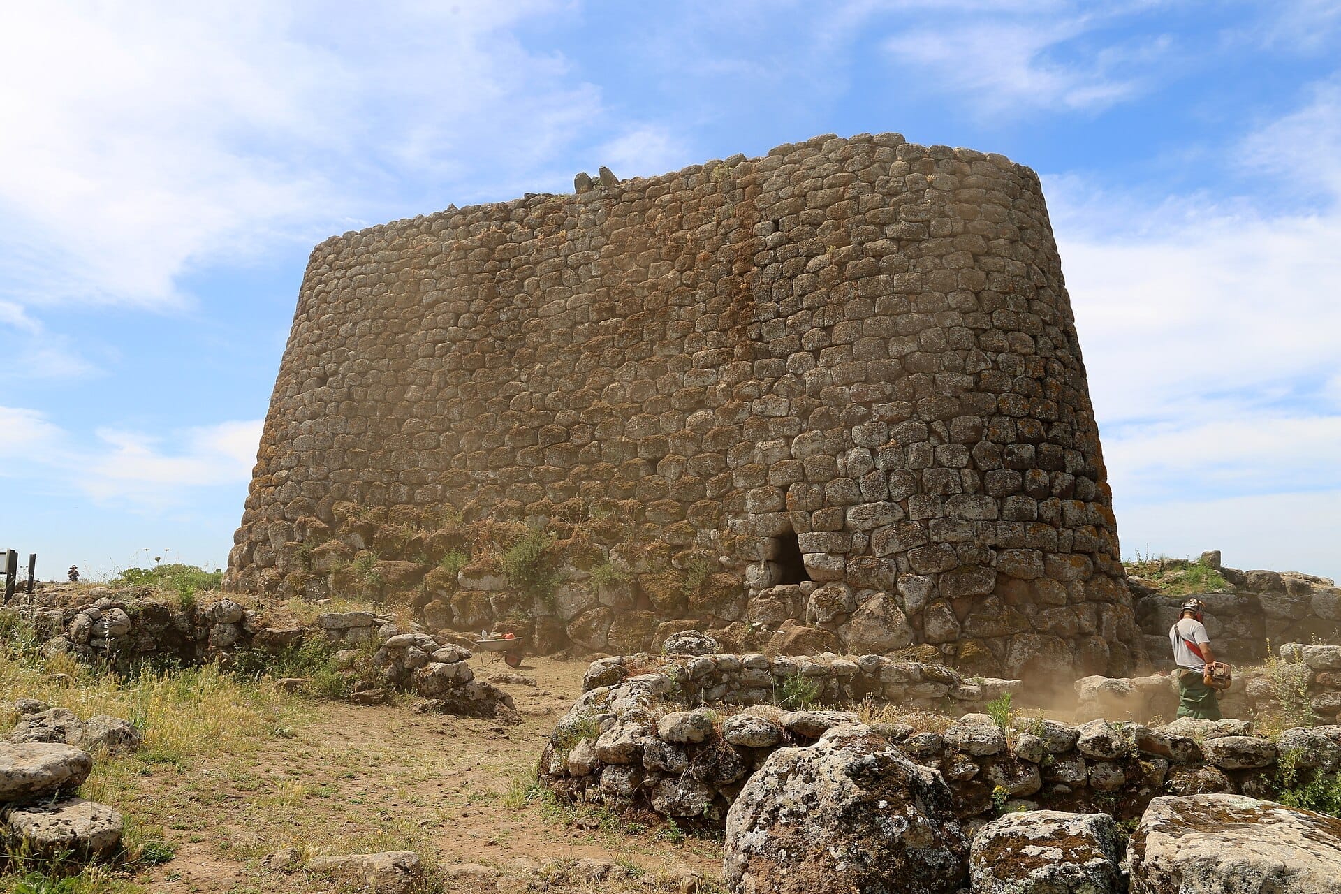 Der massive Steinturm des Nuraghe Losa erhebt sich über verstreute Ruinen und wildes Gras unter einem strahlend blauen Himmel in Sardinien.