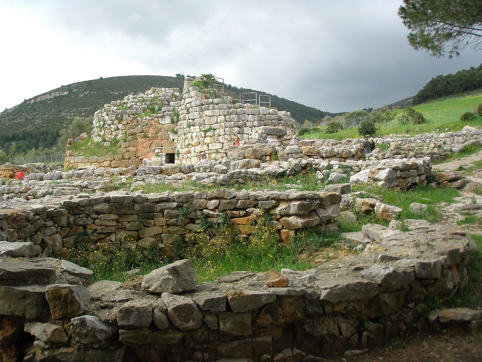 Steinruinen von Nuraghe Palmavera mit kreisförmigen Hütten und einem zentralen Turm, vor grünen Hügeln unter bewölktem Himmel in der Nähe von Alghero.