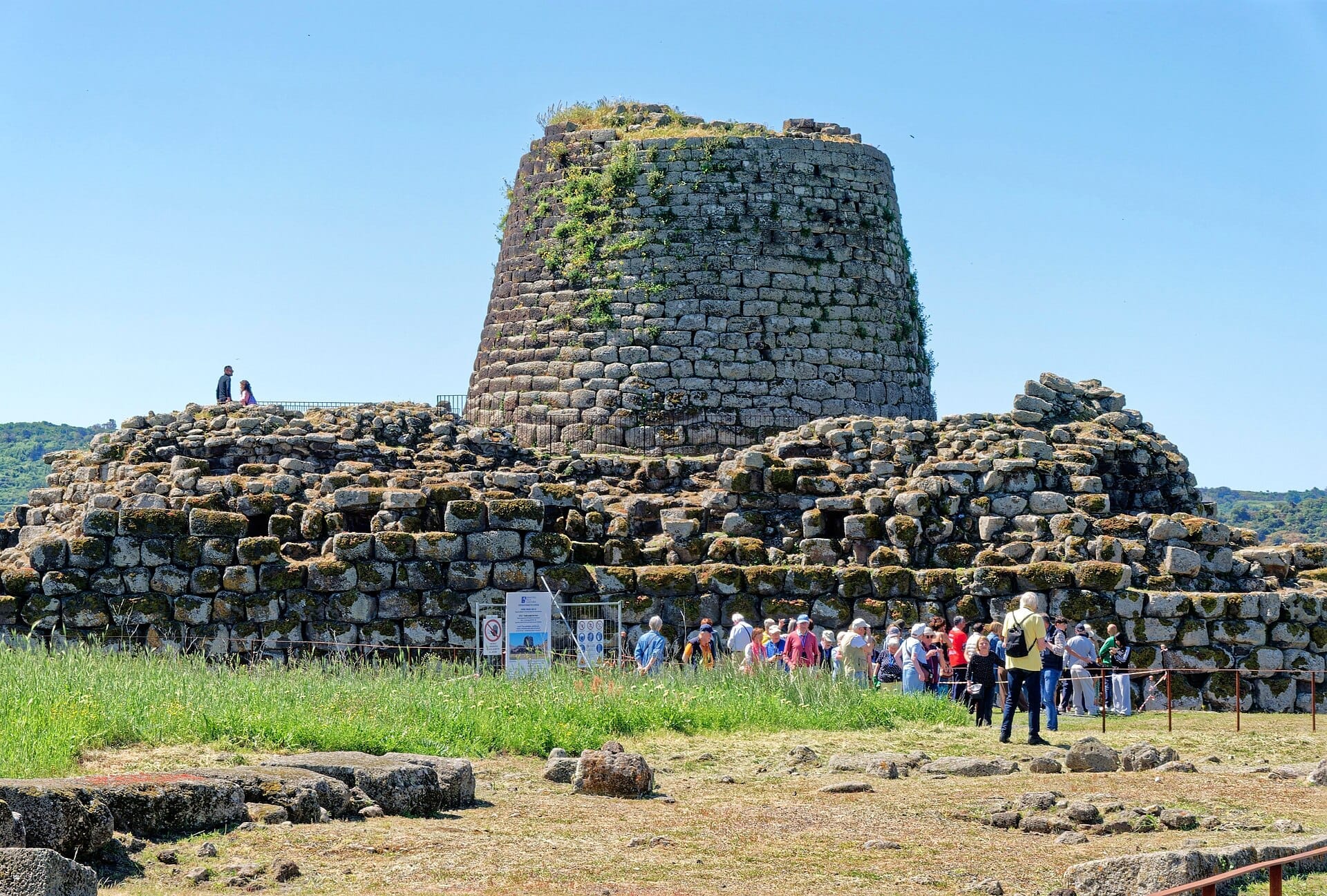 Die Steinmauern und der zentrale Turm des Nuraghe Santu Antine ragen über grasbewachsene Ruinen empor, während Besuchergruppen an einem sonnigen Tag das Gelände erkunden.