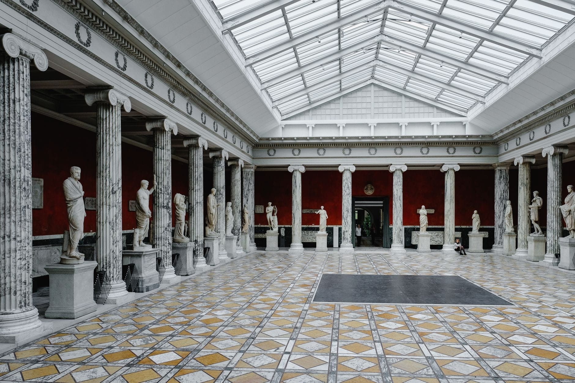 Ampia sala in marmo con soffitto a lucernario, fiancheggiata da statue antiche e colonne di marmo, con pavimento a motivi geometrici e vivaci pareti rosse alla Ny Carlsberg Glyptotek.