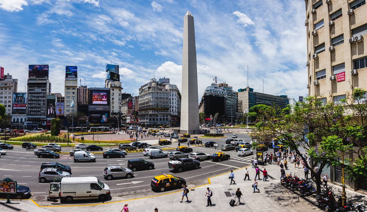 ภาพมุมกว้างกลางวันของ Obelisco de Buenos Aires ตั้งอยู่กลางสี่แยกใหญ่ รายล้อมด้วยอาคาร รถยนต์ และคนเดินเท้า ท้องฟ้าสีฟ้าสดพร้อมเมฆขาว