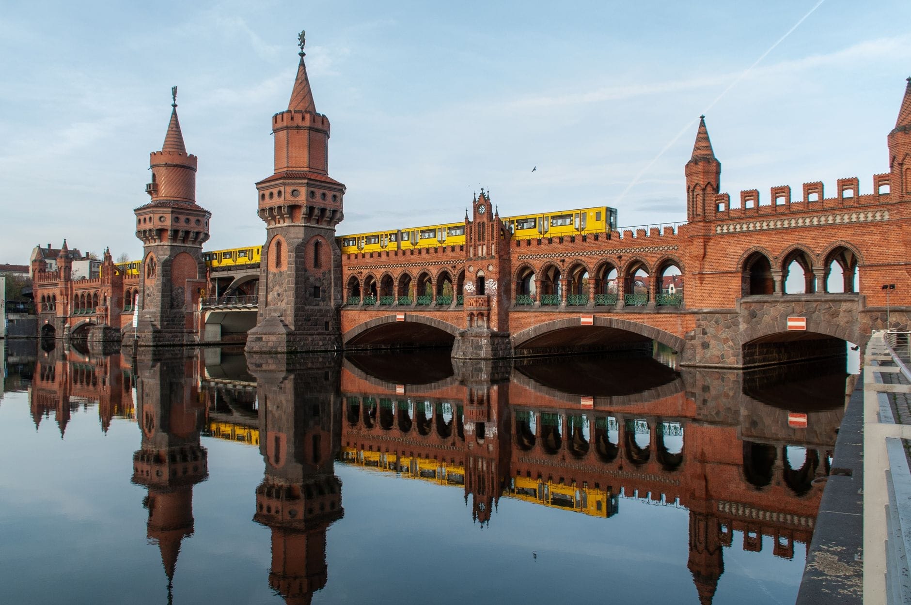 Vista panoramica dell'Oberbaumbrücke con le sue torri e arcate neo-gotiche in mattoni riflesse nelle acque calme dello Sprea, con un treno giallo della U-Bahn che attraversa il ponte sotto un cielo luminoso.