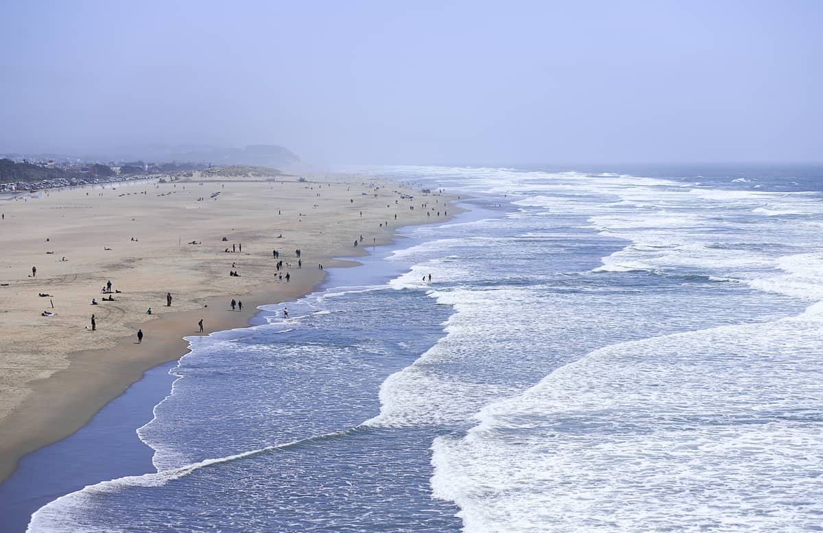Vista panoramica di Ocean Beach a San Francisco: le onde si infrangono lungo la lunga riva sabbiosa e molte persone si godono la spiaggia sotto un cielo azzurro velato.