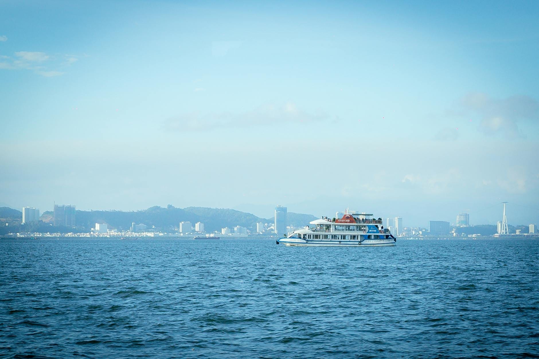 Uno yacht a più ponti naviga sulle acque calme e azzurre della baia di Pattaya, con lo skyline della città e le colline visibili sullo sfondo sotto un cielo limpido.