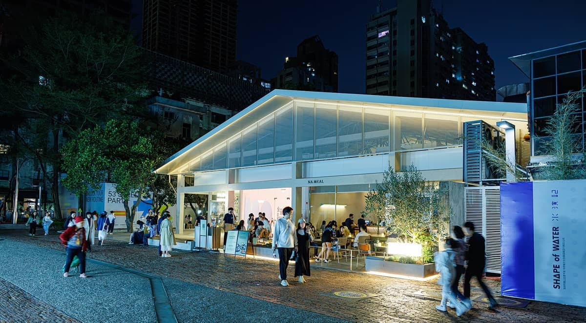 Visitantes passam por um moderno café com fachada de vidro à noite no OCT Loft Creative Culture Park, rodeado de edifícios urbanos e iluminação suave.