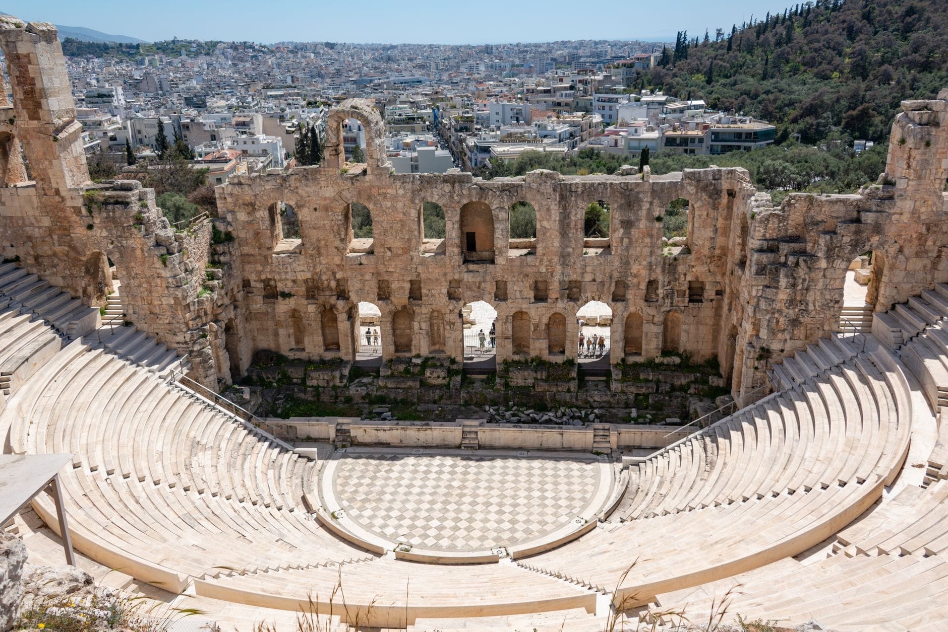 Vista grandangolare dell'Odeon di Erode Attico con le gradinate in pietra, la parete scenica ad archi e il panorama di Atene sotto una luce diurna intensa.