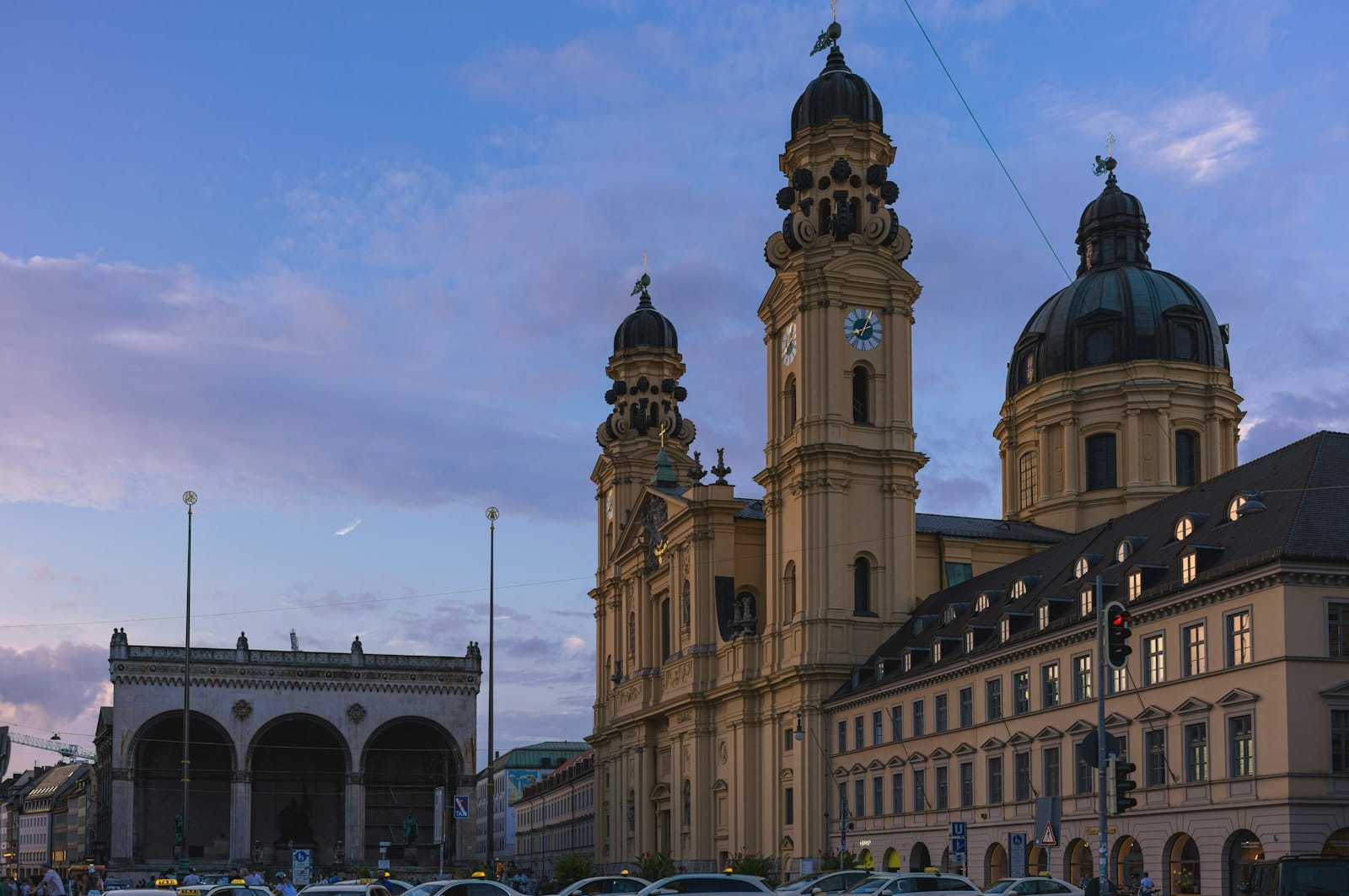 Odeonsplatz ในมิวนิกยามพลบค่ำ เห็นโบสถ์ Theatinerkirche และ Feldherrnhalle โอบล้อมด้วยสถาปัตยกรรมเก่าแก่อบอุ่น ผู้คนเดินคึกคัก