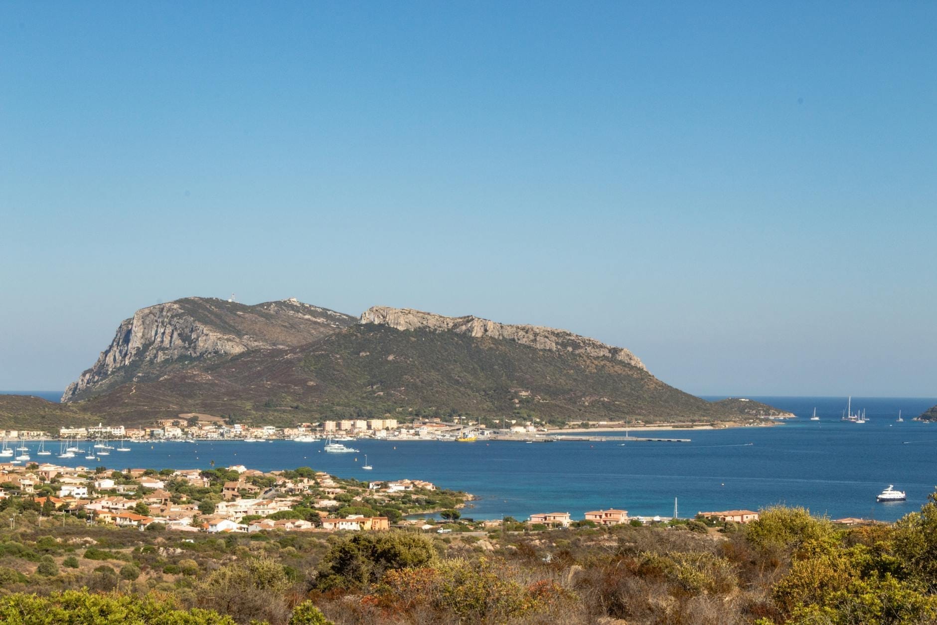 Panoramablick auf Olbia mit Meer, Segelbooten, Küstenort und einem markanten Hügel im Hintergrund unter strahlend blauem Himmel.