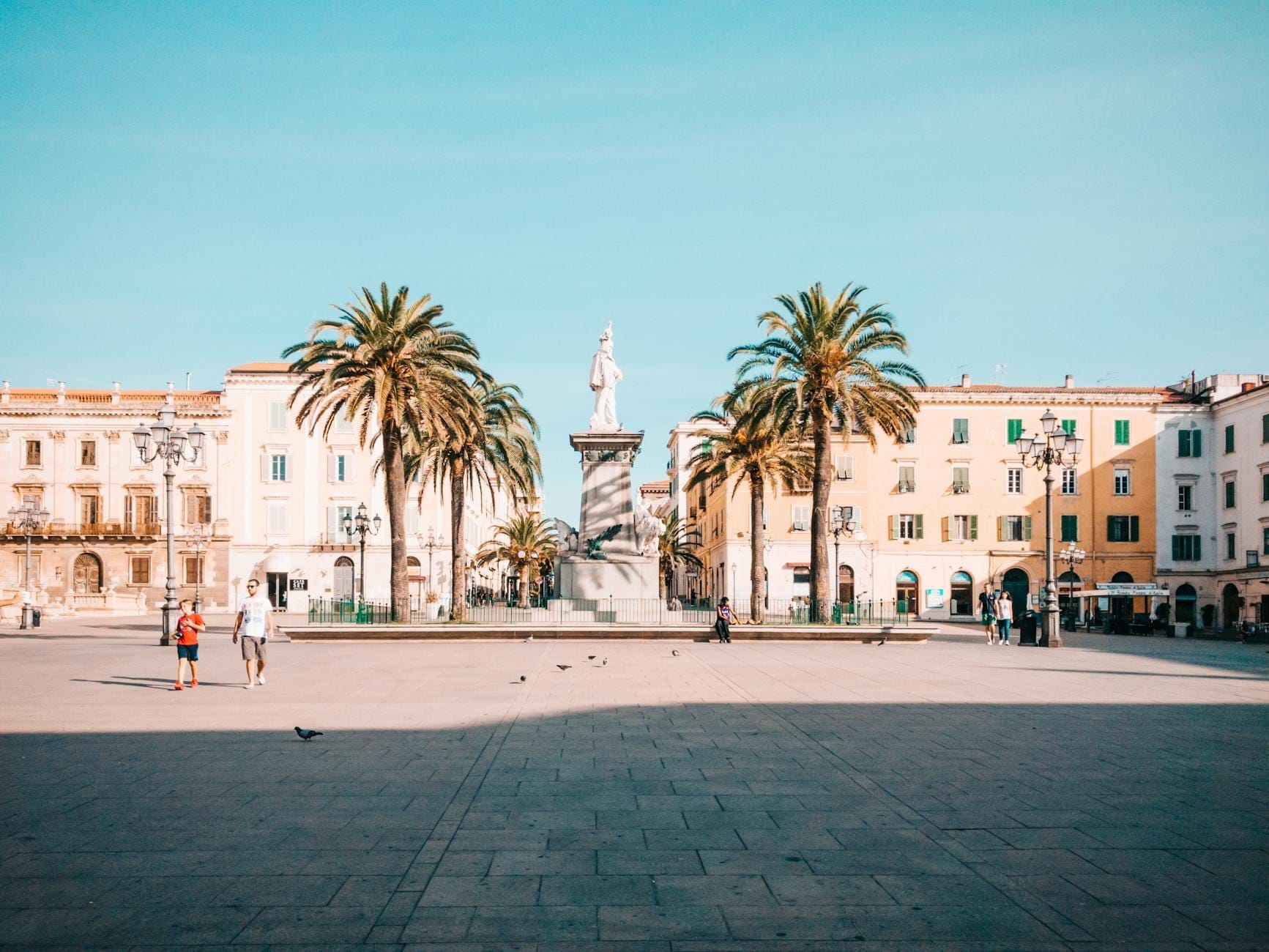 Weiter sonniger Platz auf Sardinien mit Palmen, bunten Fassaden und einer zentralen Statue, die angenehmes Wetter und eine entspannte Atmosphäre vermitteln.