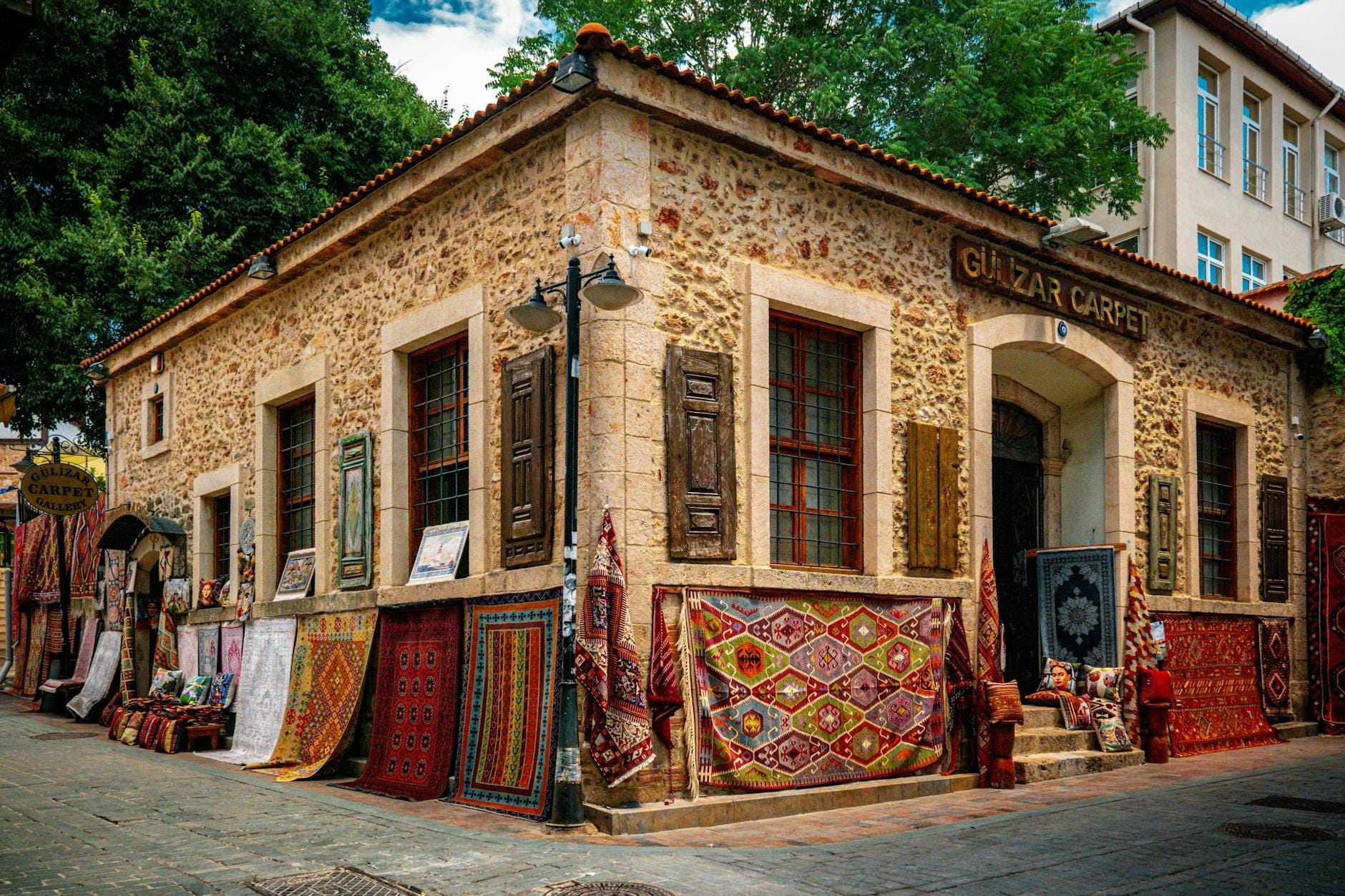 Angolo di un antico edificio in pietra nel quartiere del bazar di Antalya, con tappeti colorati, kilim e bancarelle del mercato sui vicoli acciottolati, circondato da architetture storiche.