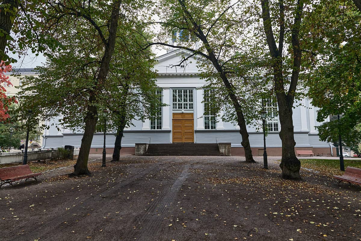 Vista frontal da Igreja Antiga de Helsinque (Vanha Kirkko) com sua fachada branca neoclássica, grandes portas de madeira e árvores num parque tranquilo no outono.