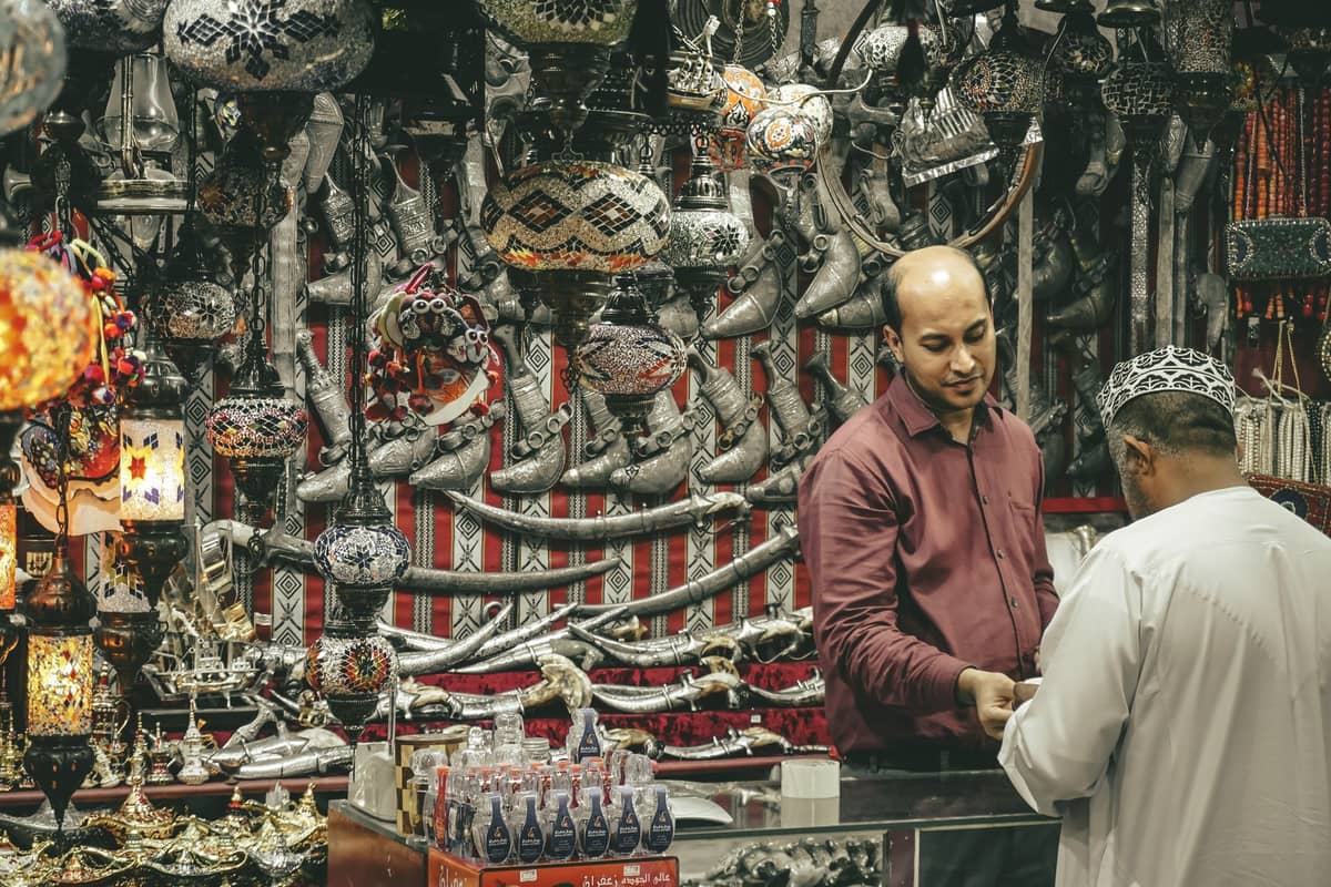 Ein belebter Stand in einem Souk im Nahen Osten mit kunstvollen Lampen, gebogenen Dolchen und traditionellen Waren, auf dem zwei Männer ein Handelsgeschäft abwickeln.