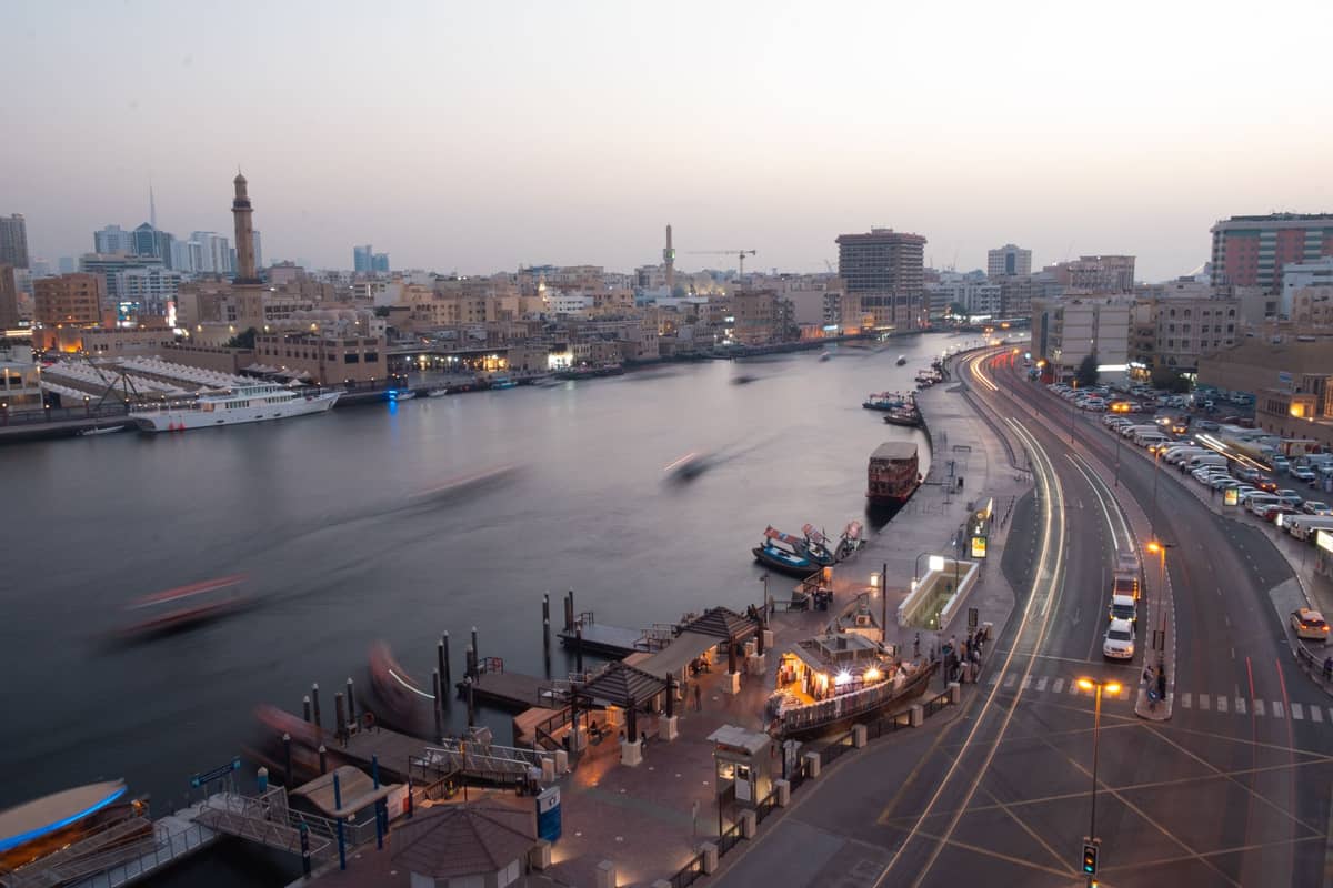 Weitblick auf den Dubai Creek mit traditionellen Holzbooten, Stadtstraßen und historischen Gebäuden entlang beider Ufer bei Sonnenuntergang.