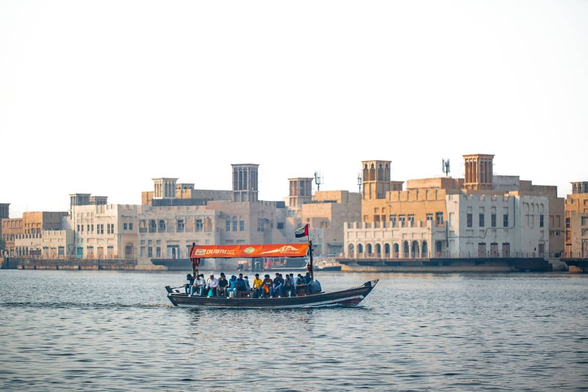 Ein hölzernes Abra-Fährboot mit Passagieren überquert den Dubai Creek, mit traditionellen Windturm-Gebäuden im Hintergrund unter klarem Himmel.