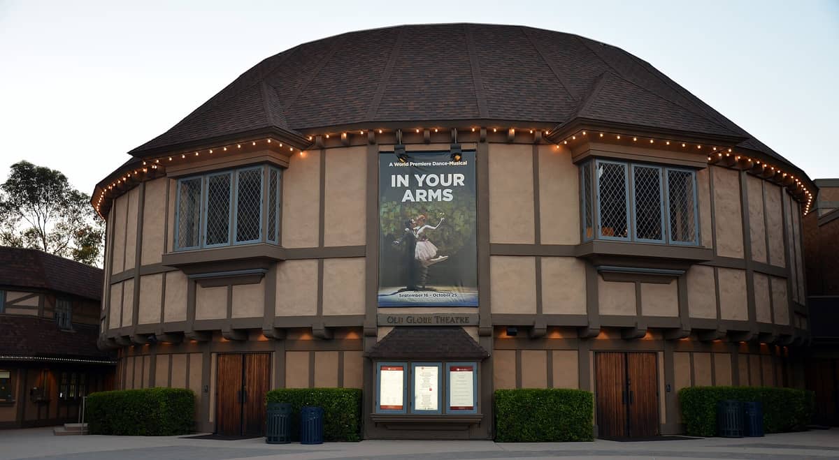 Vista panorámica del Old Globe Theatre en Balboa Park, San Diego, con su exterior de estilo Tudor, su forma hexagonal y la entrada principal bajo el cartel del teatro.