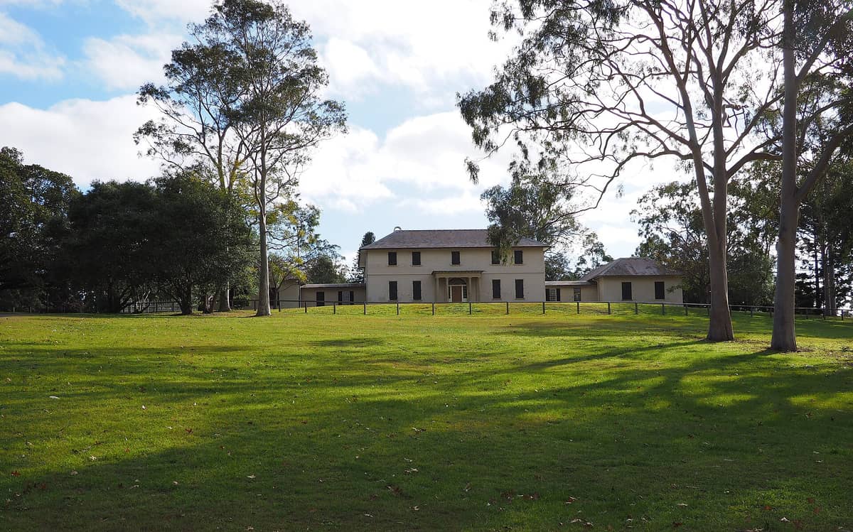 La Old Government House en Parramatta Park, un edificio colonial georgiano temprano rodeado de amplios jardines verdes, altos eucaliptos y un cielo azul de fondo.