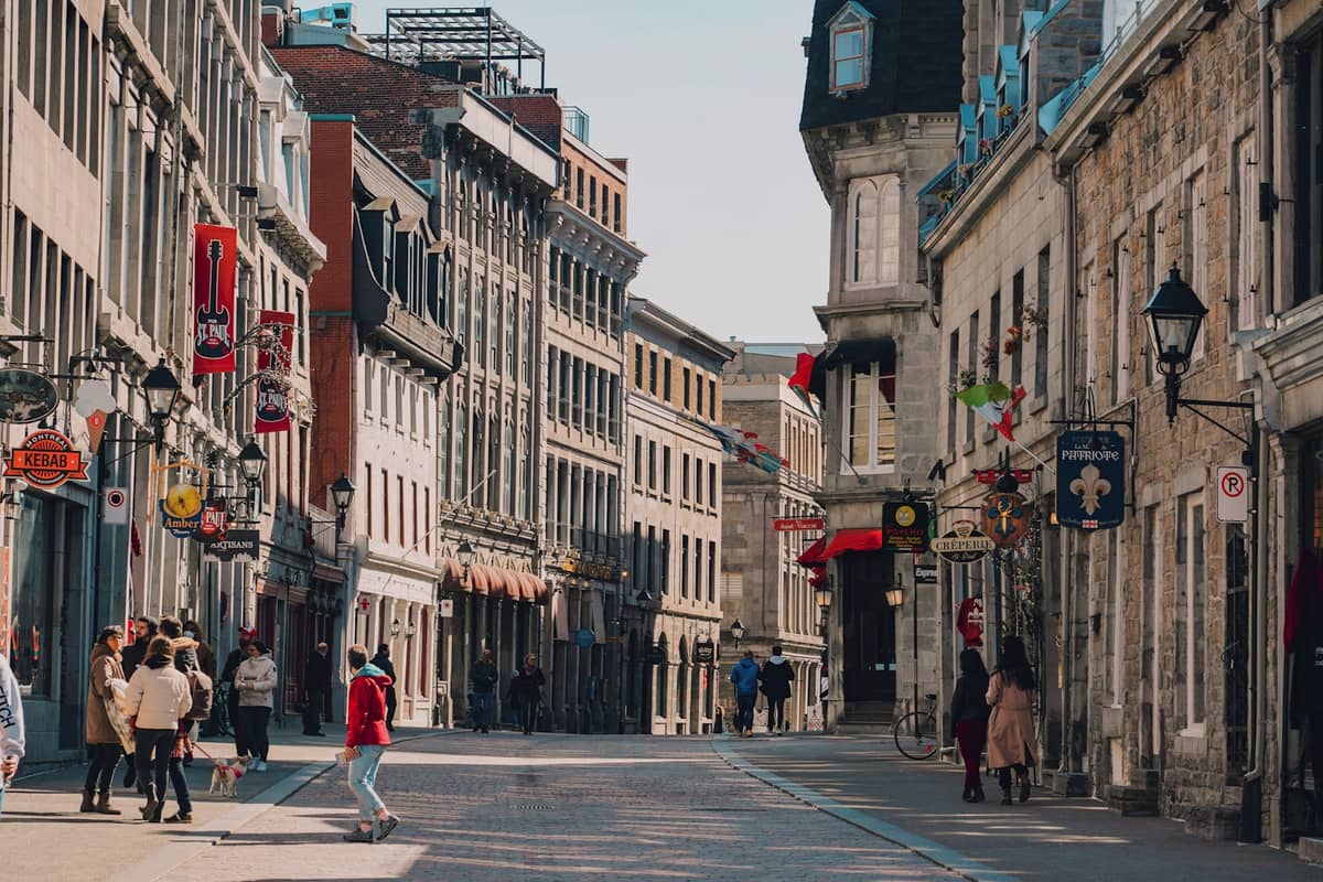 Eine belebte Straße in Alt-Montreal mit historischen Steinhäusern, französischen Schildern, Boutiquen und Menschen auf dem Kopfsteinpflaster.