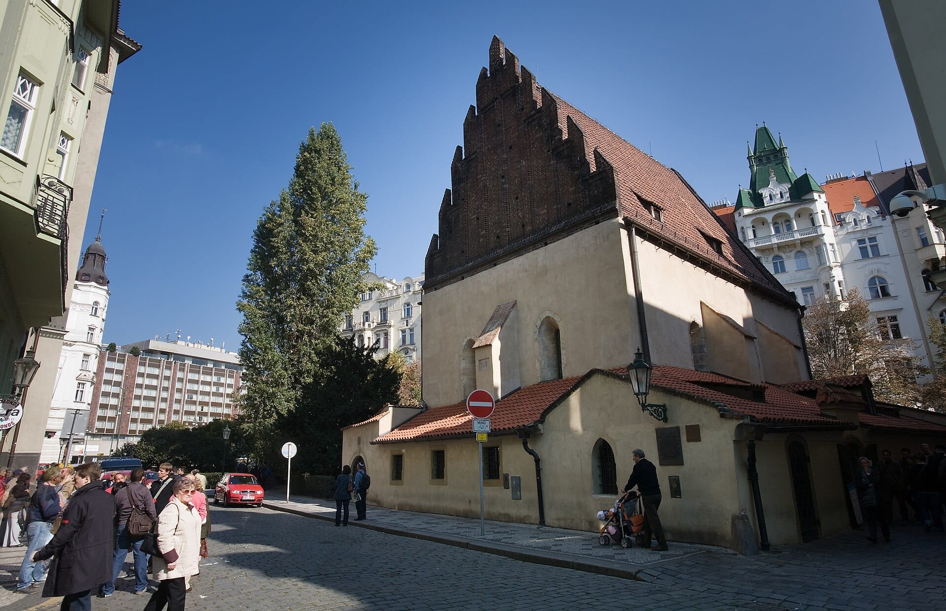 Widok zewnętrzny Staronowej Synagogi w Pradze z charakterystycznym gotyckim ceglanym szczytem i ochrowo-żółtymi ścianami pod bezchmurnym błękitnym niebem.