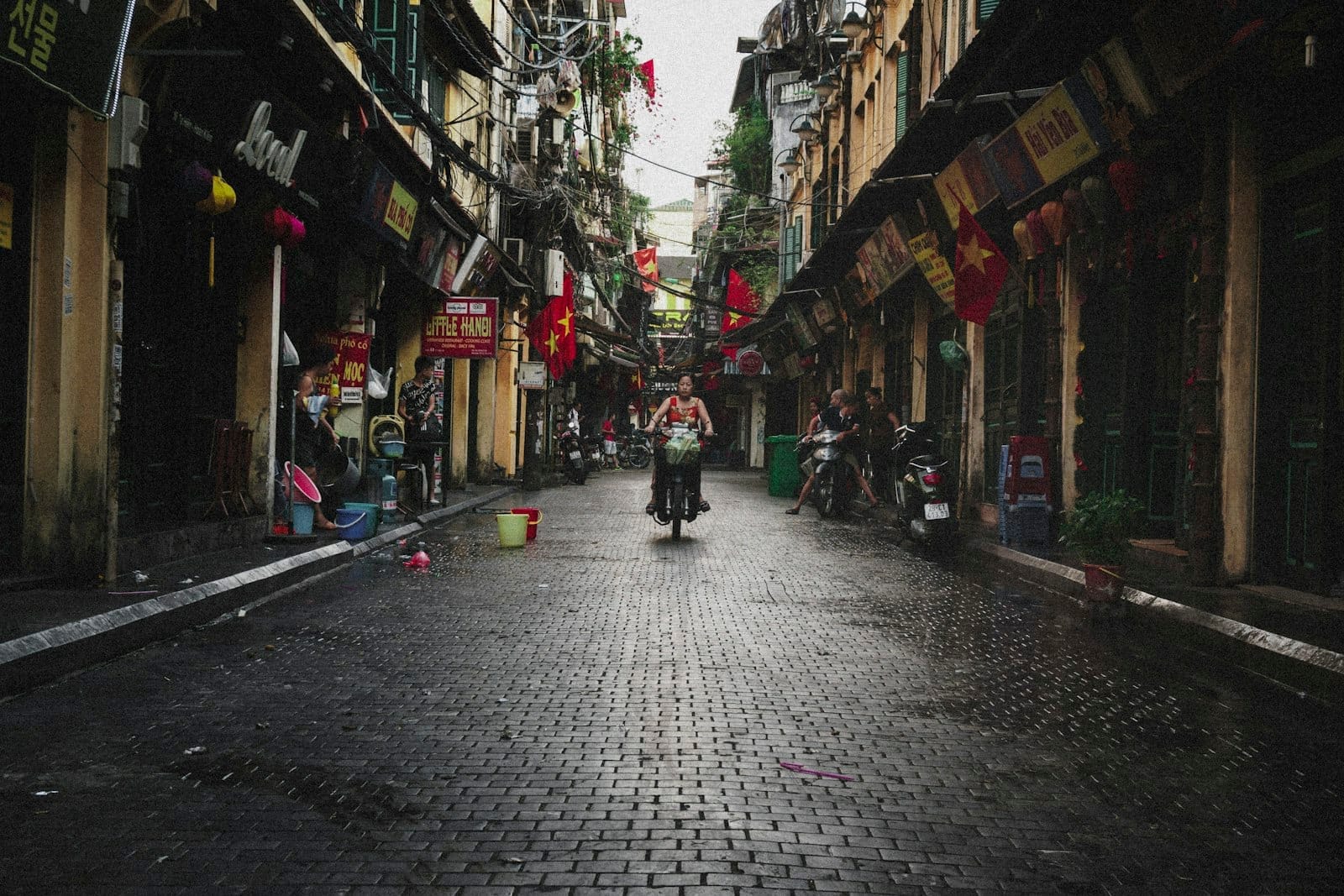 A narrow street in Hanoi's Old Quarter, lined with old colonial buildings, Vietnamese flags, and lanterns, with a person riding a bicycle down the wet cobblestone street.