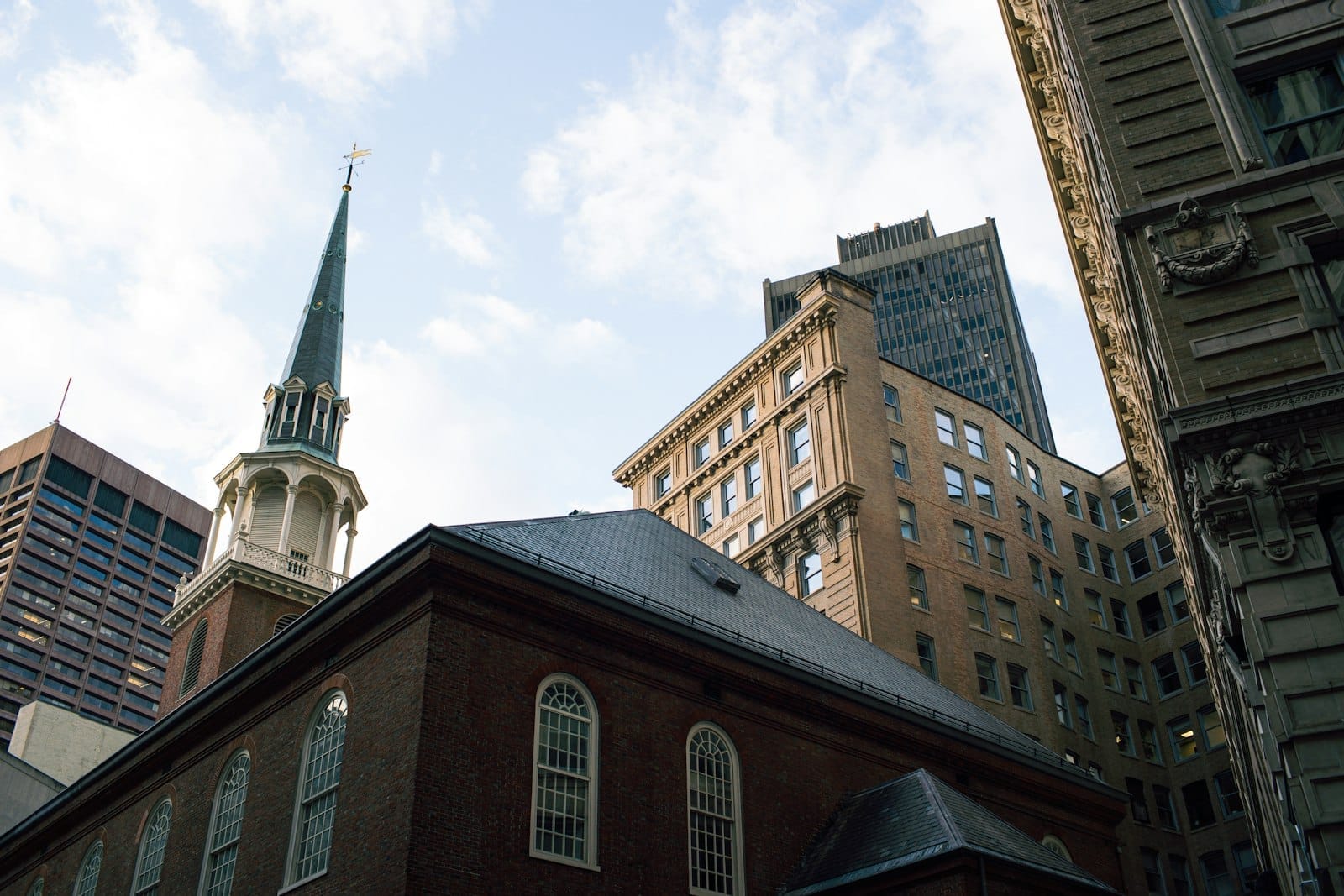 L'esterno in mattoni e il campanile dell'Old South Meeting House circondati da edifici moderni e storici del centro di Boston in una giornata parzialmente nuvolosa.