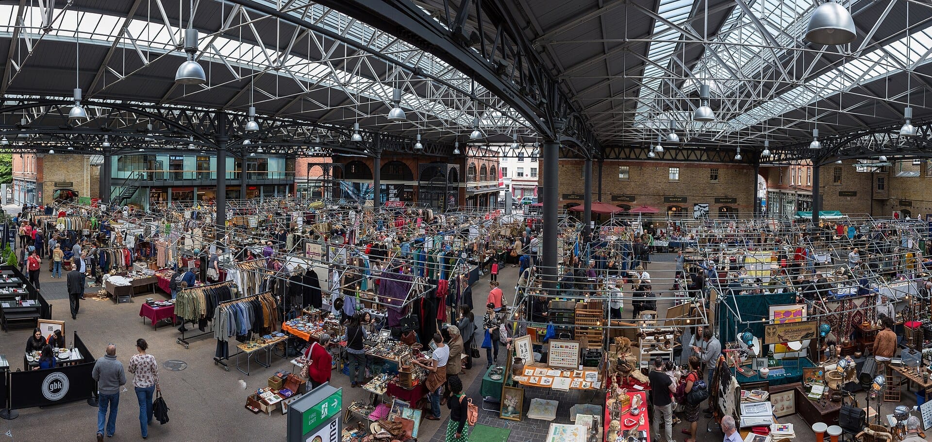 Ampia vista dell'interno dell'Old Spitalfields Market, con folla animata, venditori, abbigliamento vintage e merci sotto un tetto in ferro e vetro.