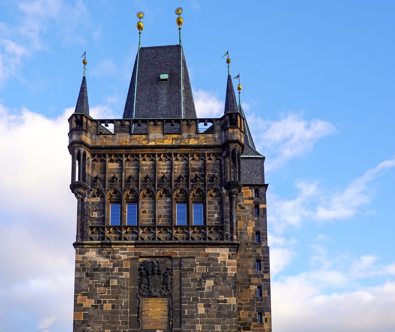 Le guglie gotiche e la dettagliata facciata in pietra della Torre del Ponte della Città Vecchia a Praga si stagliano contro un cielo azzurro brillante con nuvole sparse.