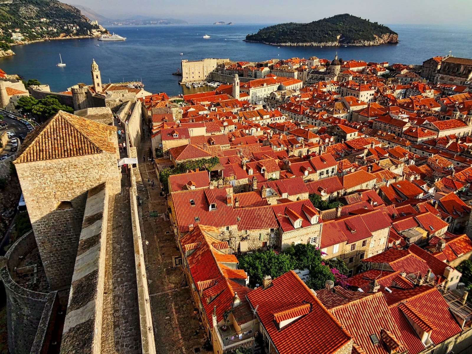 Vista aérea panorámica de la Ciudad Vieja de Dubrovnik, con sus tejados de terracota, las imponentes murallas medievales de piedra y el mar Adriático con una isla verde al fondo bajo la luz dorada del sol.